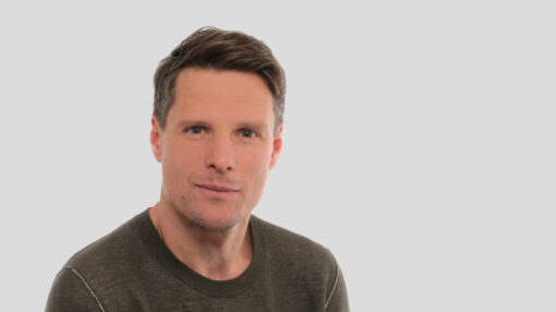 Portrait photograph of Andreas Stadelmann, a man with short brown hair, smiling slightly and wearing an olive-green jumper, against a plain light grey background